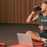 Man in gym drinking water during workout on exercise mat with laptop.