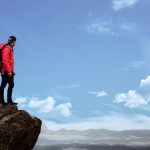 Hiker in red jacket stands on a cliff edge, overlooking a scenic mountain view under a bright blue sky.